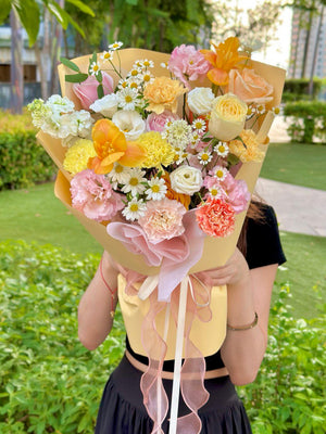 A vibrant mixed flower bouquet with orange tulips, roses, carnations, chamomile, and yellow wrapping in a greenery background.