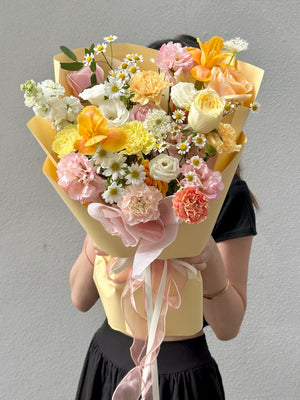 Handheld pastel-themed bouquet featuring yellow, peach, and pink blooms with a long pink ribbon.