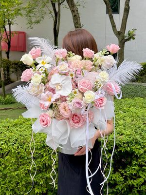 A person outdoors holding a large, elaborate floral bouquet. The bouquet features pink and white roses, Pink Hydrangea, white orchids, white daisies, white feathers, and white ribbon streamers wrapped in sheer white paper.
