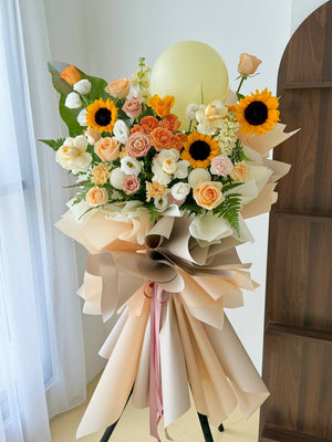A large grand opening flower stand with a bouquet of sunflowers, roses and other pastel flowers, wrapped in beige paper with a large bow and ribbon, topped with a pale yellow balloon, photographed indoors near a window and wooden cabinet.