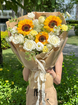 A bright and cheerful bouquet featuring sunflowers, white roses, and delicate yellow carnations wrapped in kraft and cream paper.