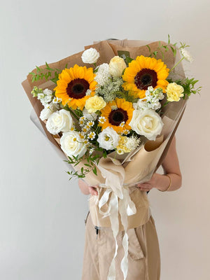 Bouquet of sunflowers and white roses wrapped in brown paper held by a person against a plain background