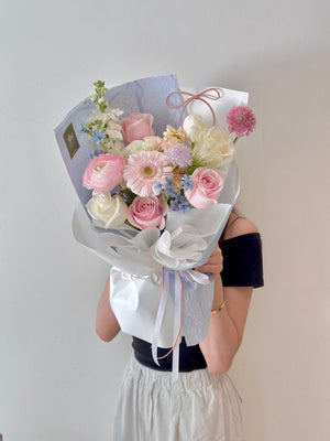 A girl in a black top smiles while holding a pink pastel bouquet with blue wrapping in her studio for her birthday.