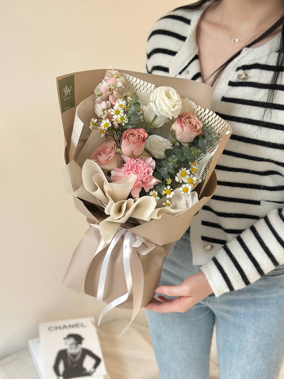 A person holding a mixed rose bouquet wrapped in beige and brown paper with green leaves and a decorative ribbon.