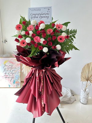 A floral arrangement featuring red roses, gerbera daisies, and green leaves, tied with a red ribbon and labeled with 'CONGRATULATIONS' and 'GRAND OPENING'.
