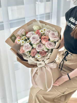 A person holding a bouquet of flowers which includes Menta roses and cappuccino roses, arranged and wrapped in brown paper with a beige ribbon.