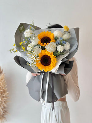 A person holding a bouquet of flowers which includes sunflowers, white roses, and a mix of other flowers with grey wrapping.