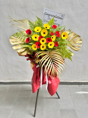 A grand opening floral arrangement featuring yellow and red Gerbera daisies, large gold Monstera leaves, a gold palm leaf, and a red ribbon, displayed on a metal stand with a 'Congratulations' message card attached.