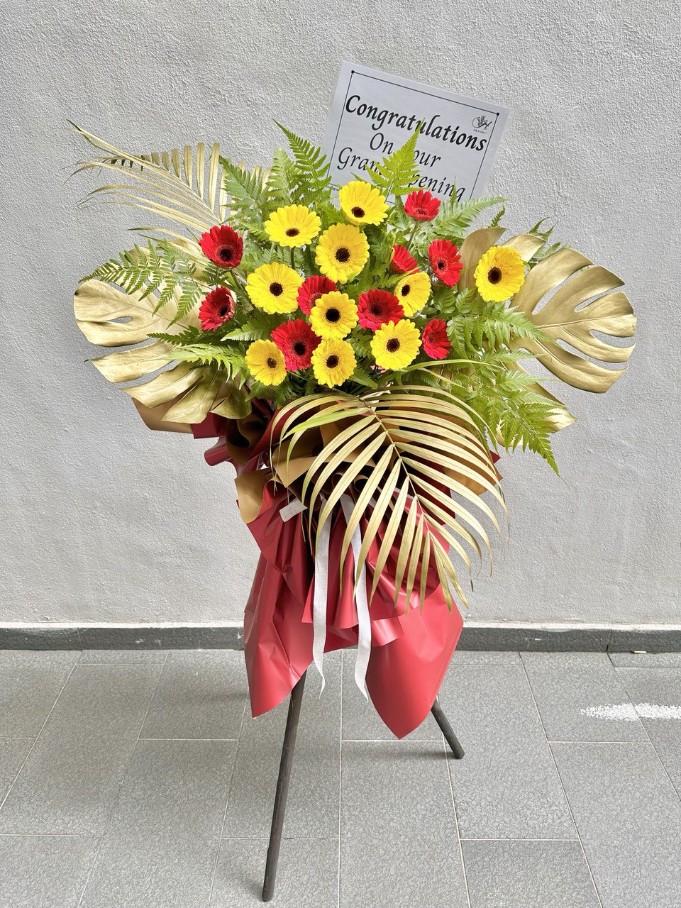 A grand opening floral arrangement featuring yellow and red Gerbera daisies, large gold Monstera leaves, a gold palm leaf, and a red ribbon, displayed on a metal stand with a 'Congratulations' message card attached.