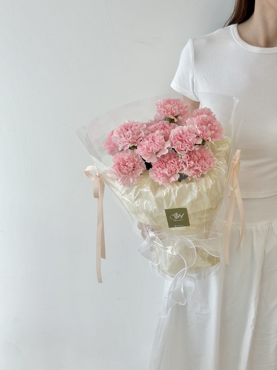 A women wearing a white top is holding a Carnation bouquet ready to surprise her mother during mother's day.