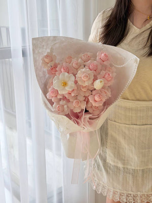 A person in a cream-colored lace dress holds a bouquet of pink kiss of fairy rose spray soap flower wrapped in sheer white paper, set against a soft, bright background with white curtains.