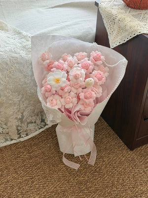 A bouquet of handmade kiss of fairy rose spray soap flower wrapped in white wrapper and a pink ribbon, placed on a brown carpeted floor next to a bed with lace bedding.