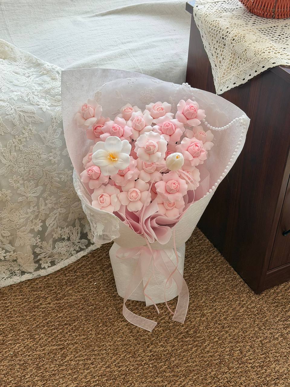 A bouquet of handmade kiss of fairy rose spray soap flower wrapped in white wrapper and a pink ribbon, placed on a brown carpeted floor next to a bed with lace bedding.