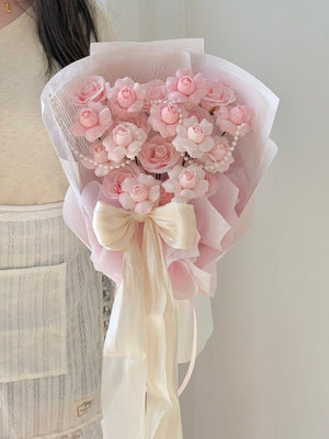 A person holding a bouquet of kiss of fairy rose spray soap flower wrapped in pale pink wrapper with a cream satin bow, against a plain light gray wall.