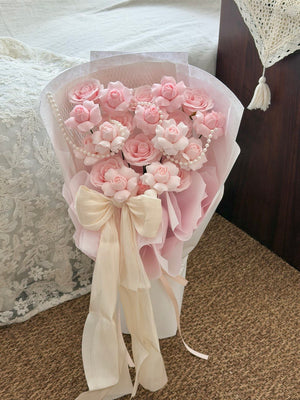 A bouquet of pink kiss of fairy rose spray soap flower roses wrapped in pale pink paper with a cream ribbon bow, standing on a carpeted floor next to a bed.