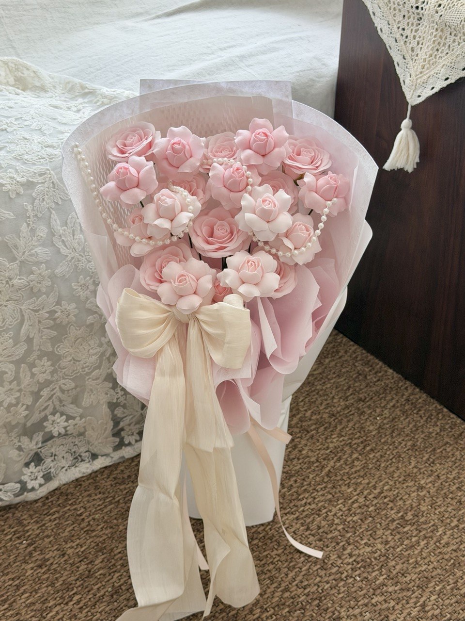 A bouquet of pink kiss of fairy rose spray soap flower roses wrapped in pale pink paper with a cream ribbon bow, standing on a carpeted floor next to a bed.