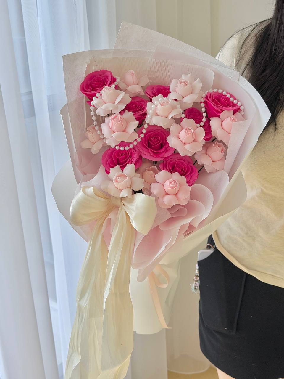 A bouquet of kiss of fairy rose spray soap flower and hot pink rose soap flower, wrapped in sheer cream paper with a cream ribbon bow, held by a person wearing a pale yellow top and black skirt.