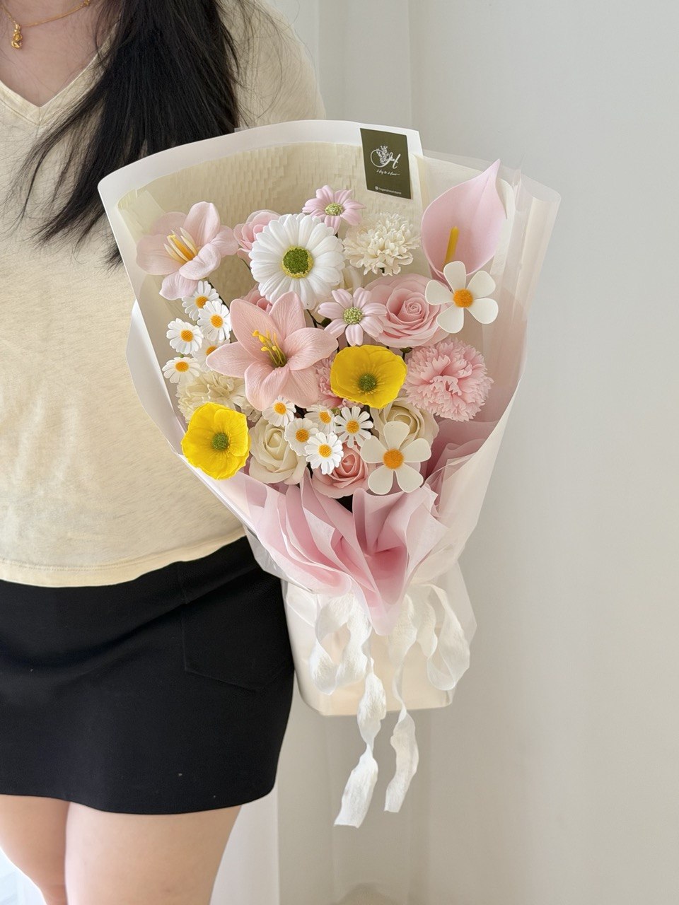 A person holding a wrapped bouquet of mixed flowers, including pink roses, white daisies, yellow flowers, and pink calla lilies, wrapped in cream paper with a pink inner layer and white ribbon.