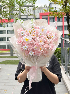 A person outdoors holding a large bouquet of kiss of fairy rose spray soap flower wrapped in sheer white paper with pink ribbons. Buildings, trees, and a railing are visible in the background.