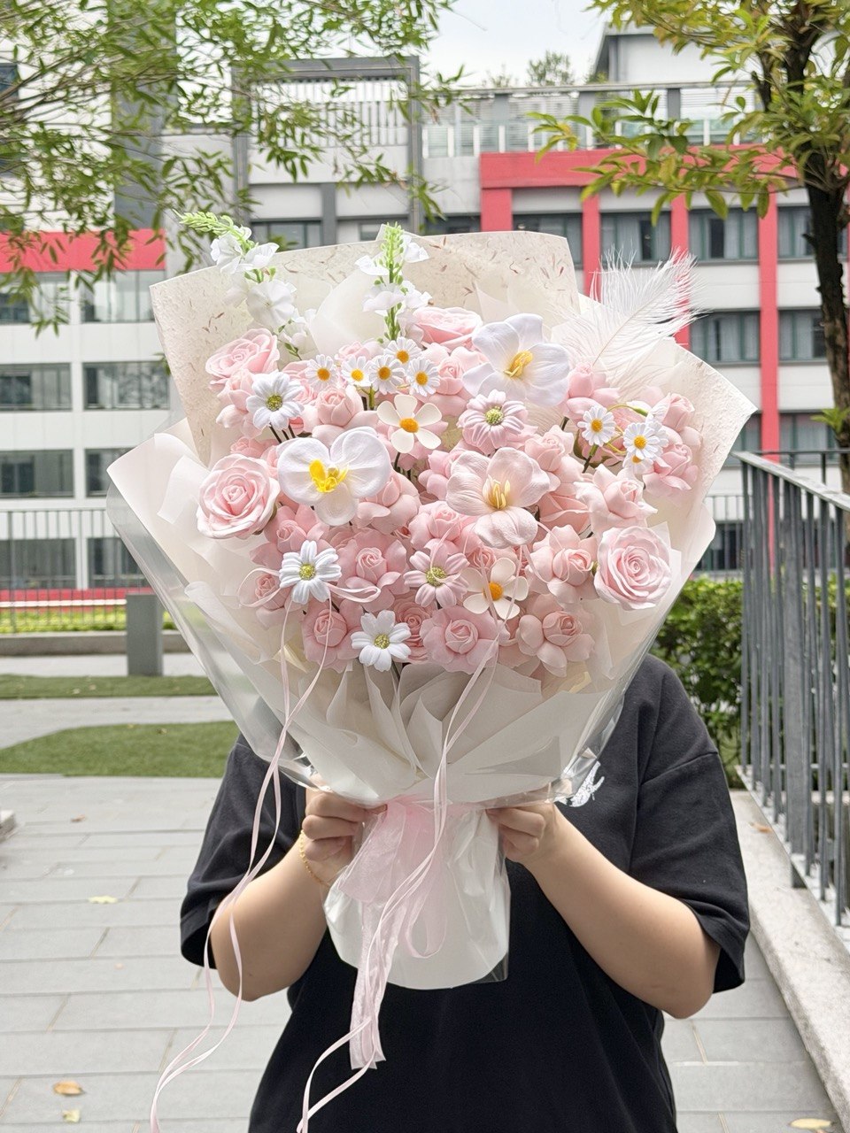 A person outdoors holding a large bouquet of kiss of fairy rose spray soap flower wrapped in sheer white paper with pink ribbons. Buildings, trees, and a railing are visible in the background.