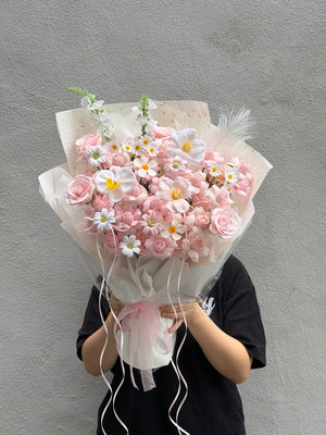 A person in a black shirt holds a large bouquet of pink kiss of fairy rose spray soap flower and white daisies wrapped in sheer white paper with pink ribbon ties, standing against a textured gray wall.