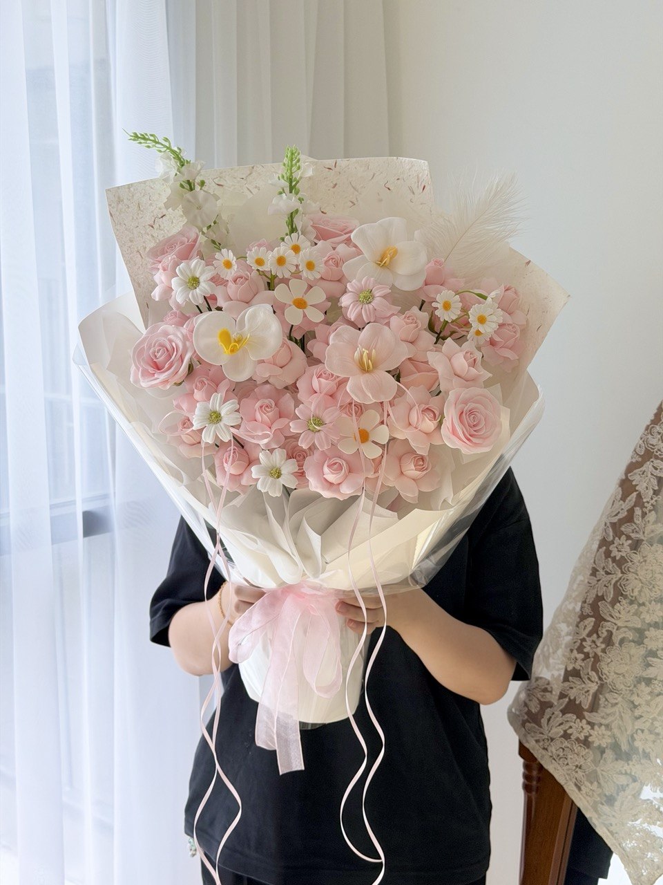 A person in a black shirt holds a large bouquet of kiss of fairy rose spray soap flower wrapped in cream paper with a pink ribbon, standing indoors near a window with sheer curtains.