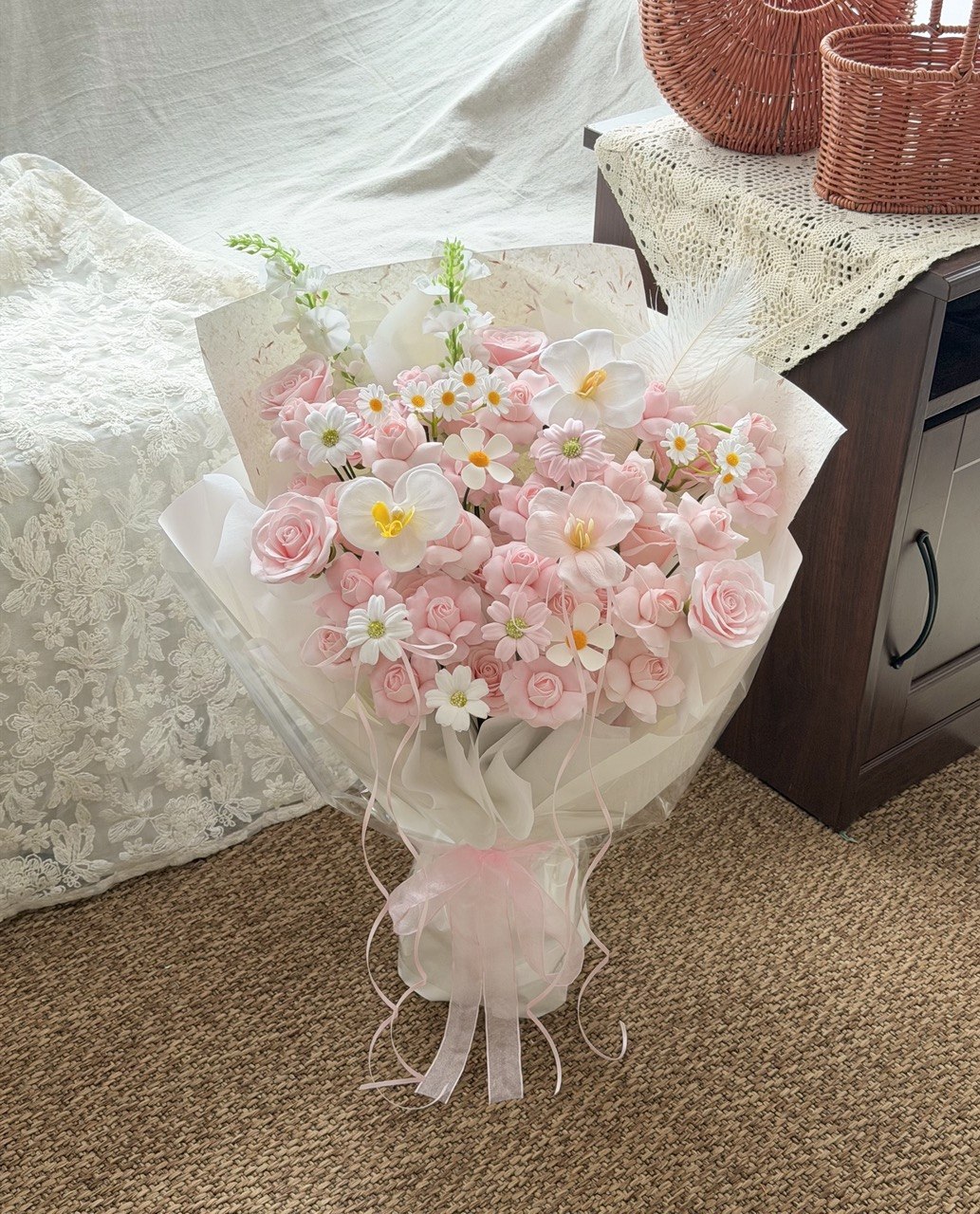 A bouquet of Kiss of fairy rose spray soap flower wrapped in sheer white paper and tied with a pink ribbon, placed on a carpeted floor next to a wooden cabinet with lace fabric.