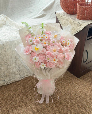 A bouquet of Kiss of fairy rose spray soap flower wrapped in sheer white paper and tied with a pink ribbon, placed on a carpeted floor next to a wooden cabinet with lace fabric.