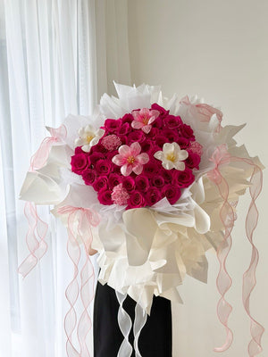 A girl is holding a heart-shaped bouquet of hot pink roses with with tulips and carnation wrapped in white ballet wrapper with pink ribbons.