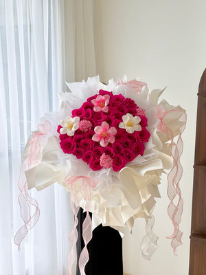 A large floral bouquet featuring heart-shaped bouquet of hot pink roses with tulips and carnation wrapped in white ballet wrapper with pink ribbons, wrapped in white and pink tissue paper with long pink ribbons, displayed on a black stand.