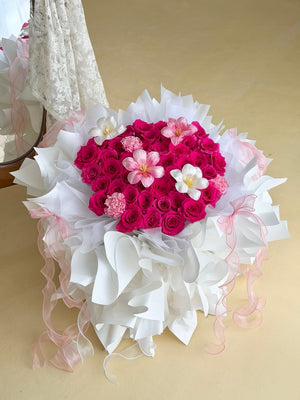 A heart-shaped bouquet of hot pink roses with tulips and carnation wrapped in white ballet wrapper with pink ribbons, set against a plain beige background.