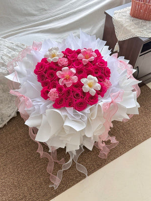 A heart-shaped bouquet of hot pink roses with tulips and carnation wrapped in white ballet wrapper with pink ribbons, placed on a brown carpeted floor.