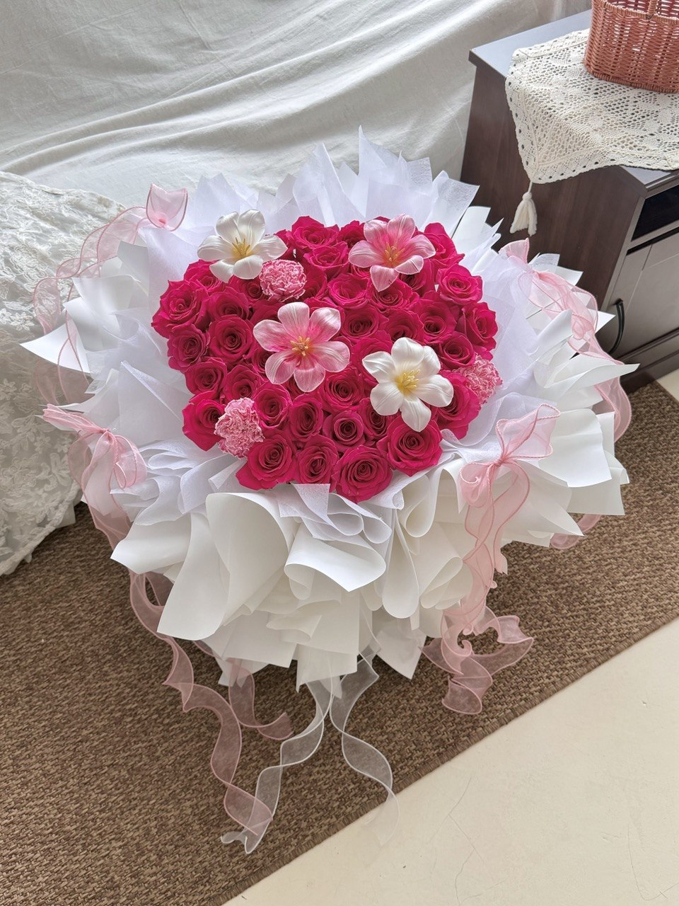 A heart-shaped bouquet of hot pink roses with tulips and carnation wrapped in white ballet wrapper with pink ribbons, placed on a brown carpeted floor.