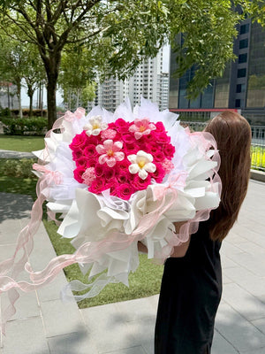 A girl is holding a heart-shaped bouquet of hot pink roses with tulips and carnation wrapped in white ballet wrapper with pink ribbons.