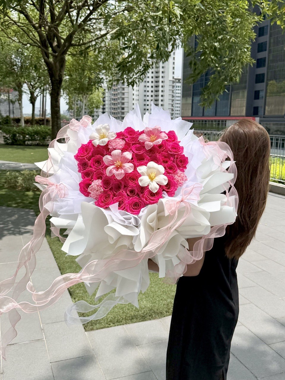 A girl is holding a heart-shaped bouquet of hot pink roses with tulips and carnation wrapped in white ballet wrapper with pink ribbons.
