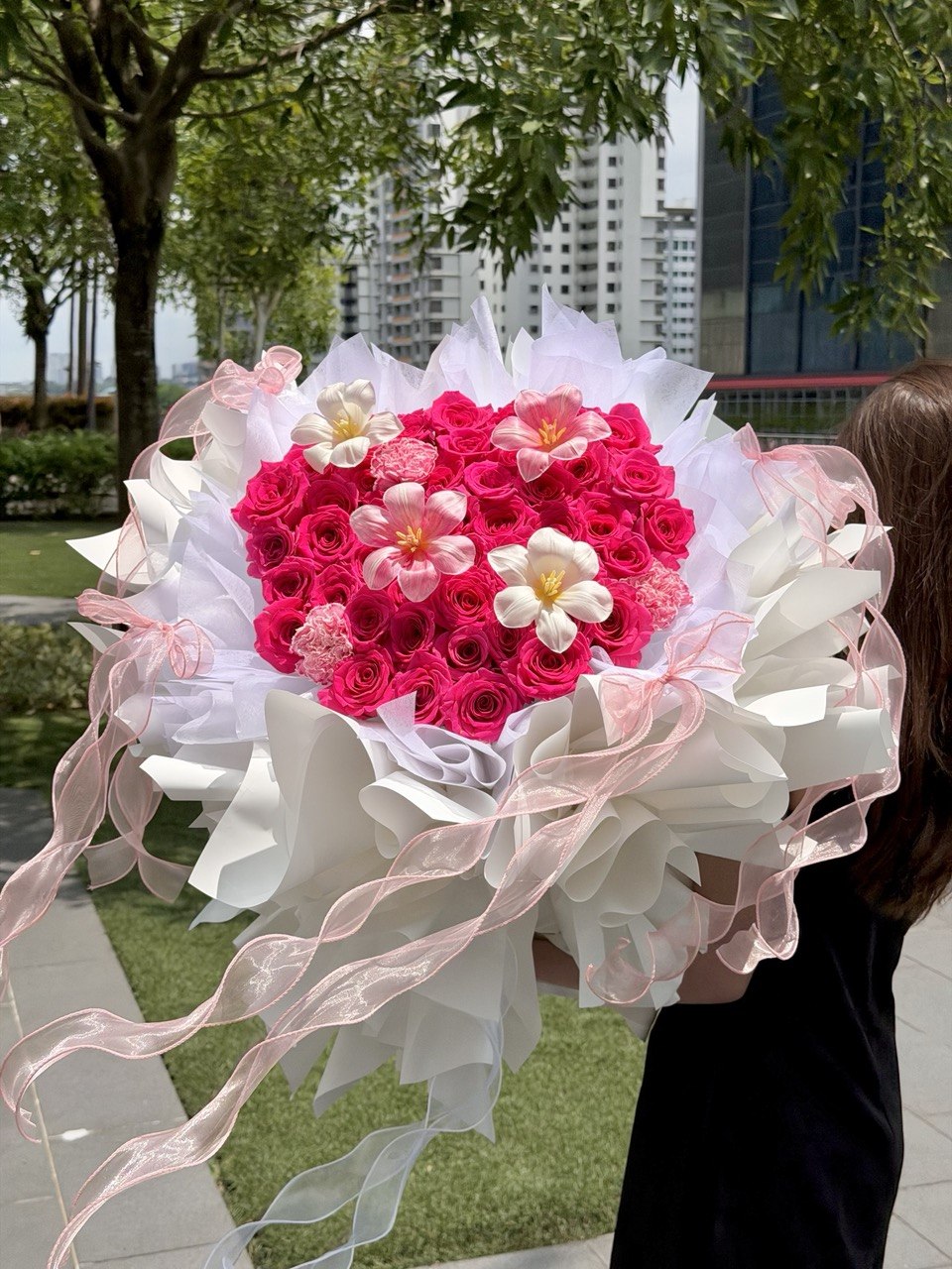 A large heart-shaped flower bouquet held by a person outdoors. The bouquet features a dense center of vibrant pink and red roses with a few white and pink accent flowers, wrapped in white paper with long pink and white ribbons.