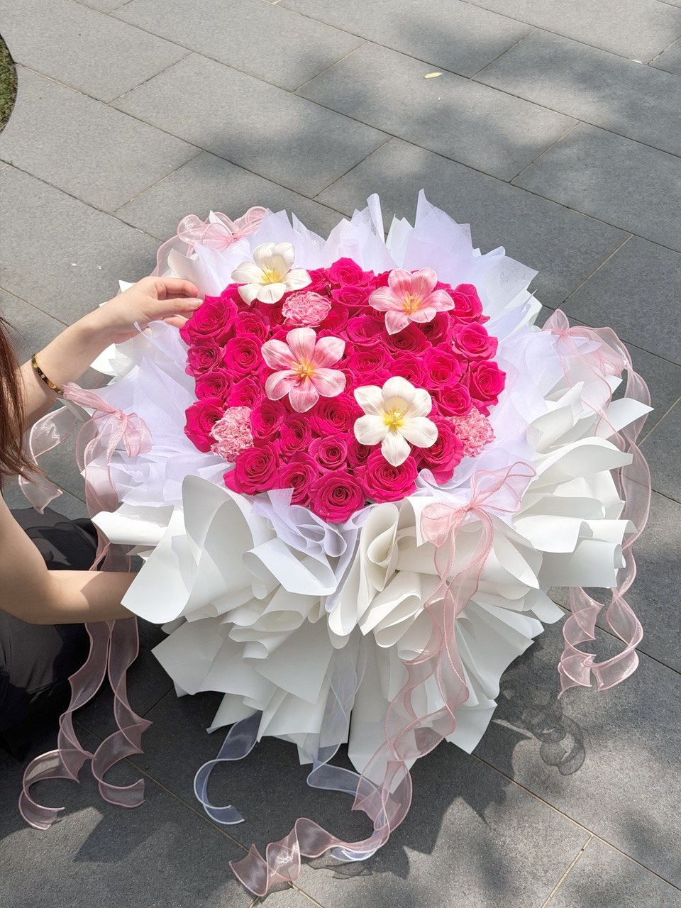A large heart-shaped flower bouquet made of bright hot pink roses with white tulip and carnation, wrapped in white ballet wrapper and pink sheer ribbons, being held by a person outdoors on a gray paved surface.