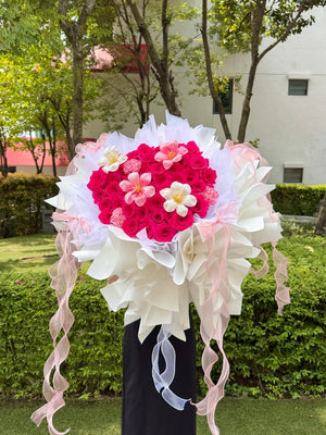 A large heart-shaped flower bouquet featuring vibrant pink roses, accented with white tulip and pink carnation, wrapped in white and pink tissue paper with long pink ribbons, displayed outdoors on a black stand.