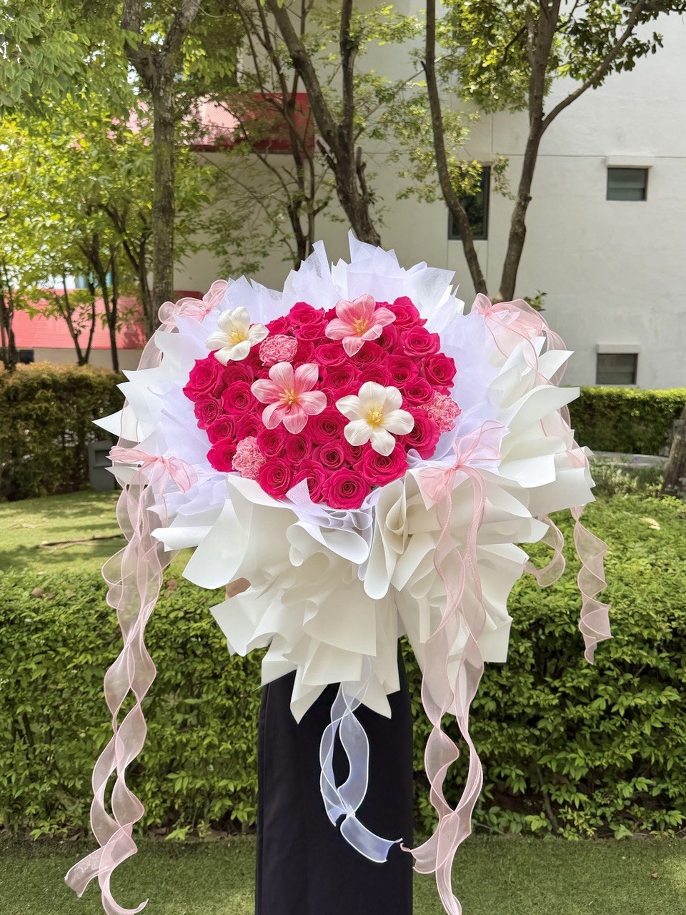 A large heart-shaped flower bouquet featuring vibrant pink roses, accented with white tulip and pink carnation, wrapped in white and pink tissue paper with long pink ribbons, displayed outdoors on a black stand.