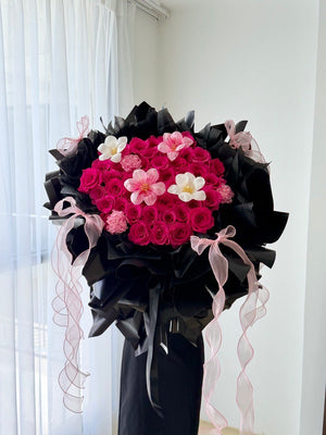 A large floral bouquet featuring hot pink roses wrapped in black paper wrapping with pink ribbon bows, displayed indoors against a white curtain and wall.
