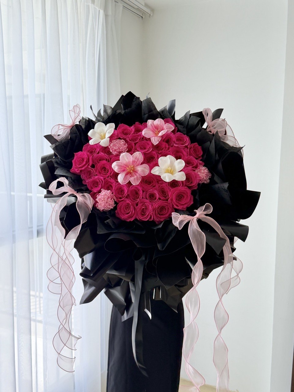 A large floral bouquet featuring hot pink roses wrapped in black paper wrapping with pink ribbon bows, displayed indoors against a white curtain and wall.
