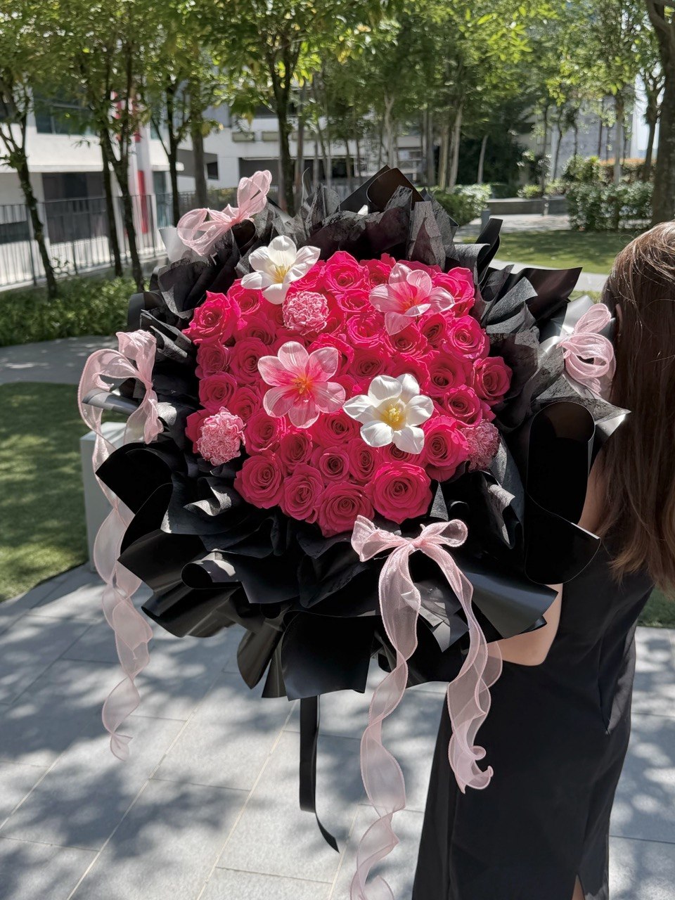 A large, vibrant flower bouquet held by a person outdoors. The bouquet features hot pink roses, tulips and carnation, wrapped in black paper with light pink ribbons tied in bows.