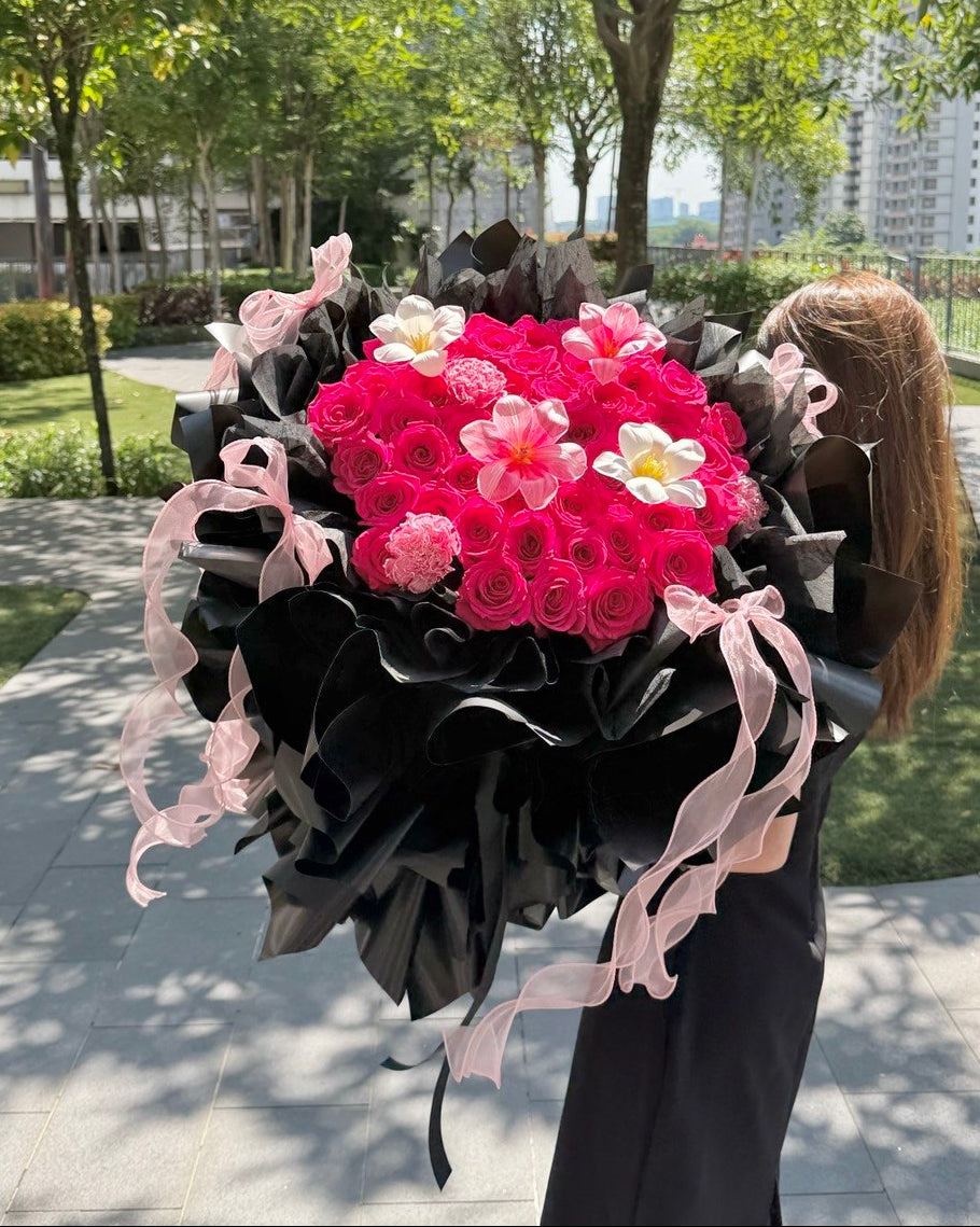 A large, vibrant flower bouquet held by a person outdoors. The bouquet features a dense cluster of bright pink roses and pink carnations, accented with white tulip and carnation, wrapped in black paper with light pink ribbons.
