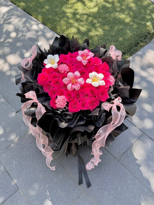 A large, vibrant flower bouquet featuring hot pink roses, accented white tulip and carnation, wrapped in dramatic black paper with sheer light pink ribbons, set on a gray paved outdoor surface.