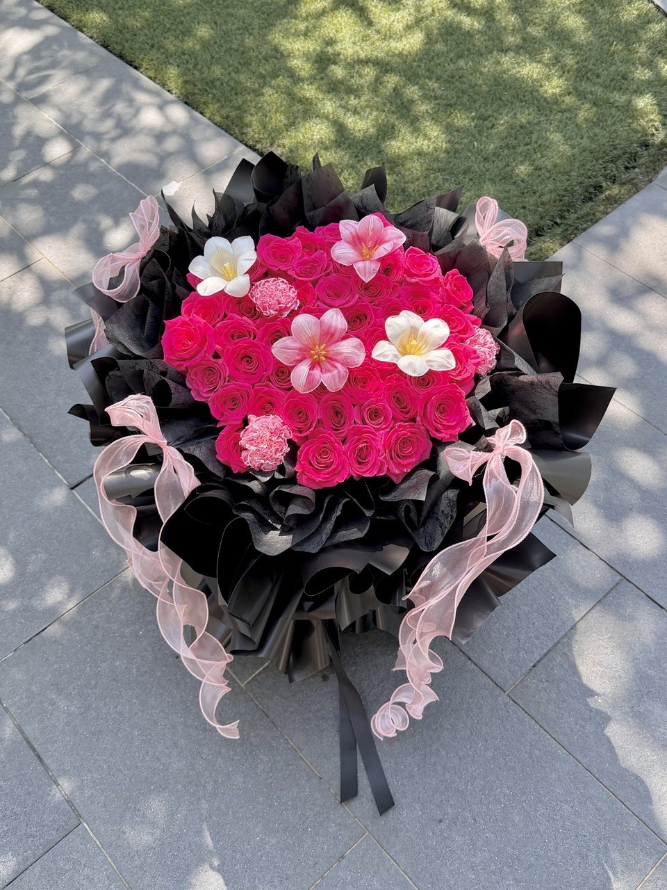 A large, vibrant flower bouquet featuring hot pink roses, accented white tulip and carnation, wrapped in dramatic black paper with sheer light pink ribbons, set on a gray paved outdoor surface.