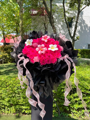A large floral bouquet featuring bright pink roses, wrapped in black paper with long sheer pink ribbons, photographed outdoors on a post with green hedges and trees in the background.