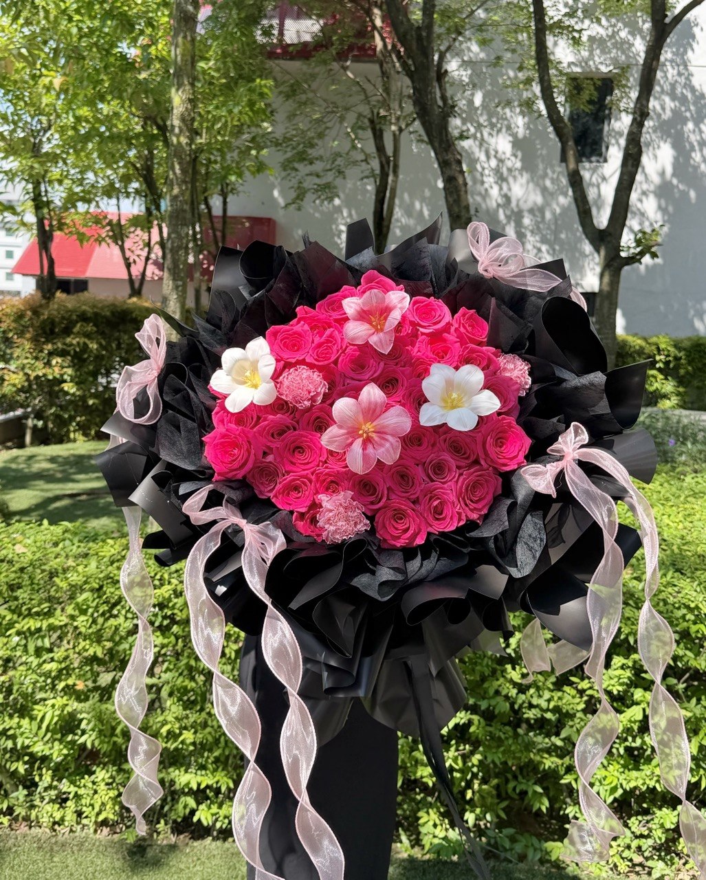 A large floral bouquet featuring bright pink roses, tulips and carnation wrapped in black paper with pink sheer ribbon tails, photographed outdoors in a garden setting.
