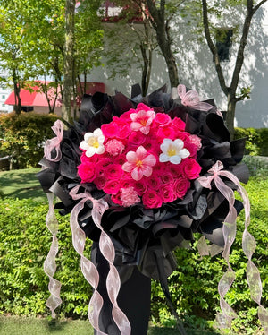 A large floral bouquet featuring bright pink roses, tulips and carnation wrapped in black paper with pink sheer ribbon tails, photographed outdoors in a garden setting.