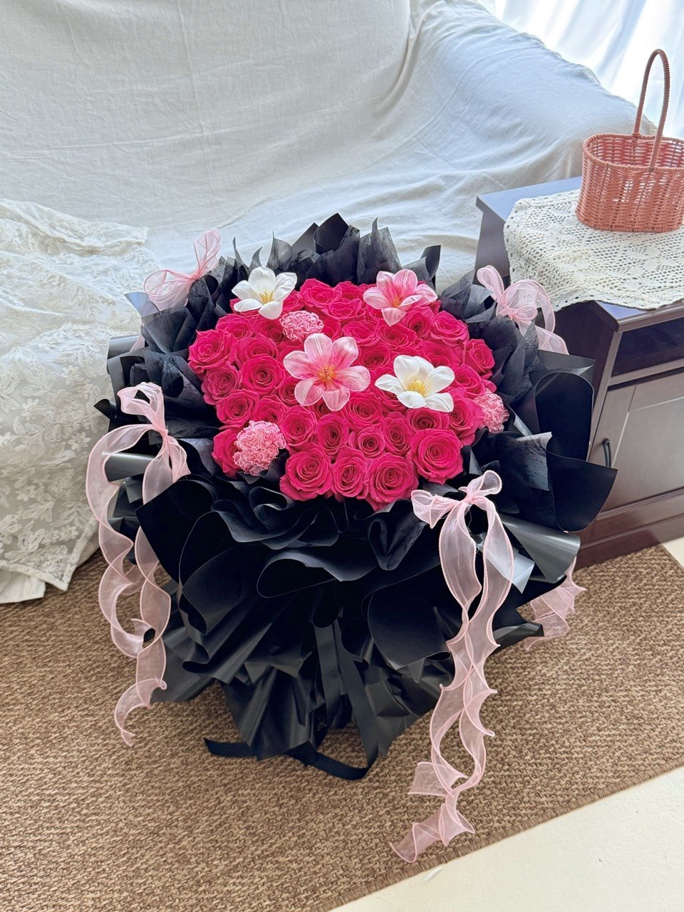 A large floral bouquet featuring bright pink roses, pink tulips, and carnations, wrapped in black paper with pink ribbon accents, placed on a carpeted floor in an indoor setting.