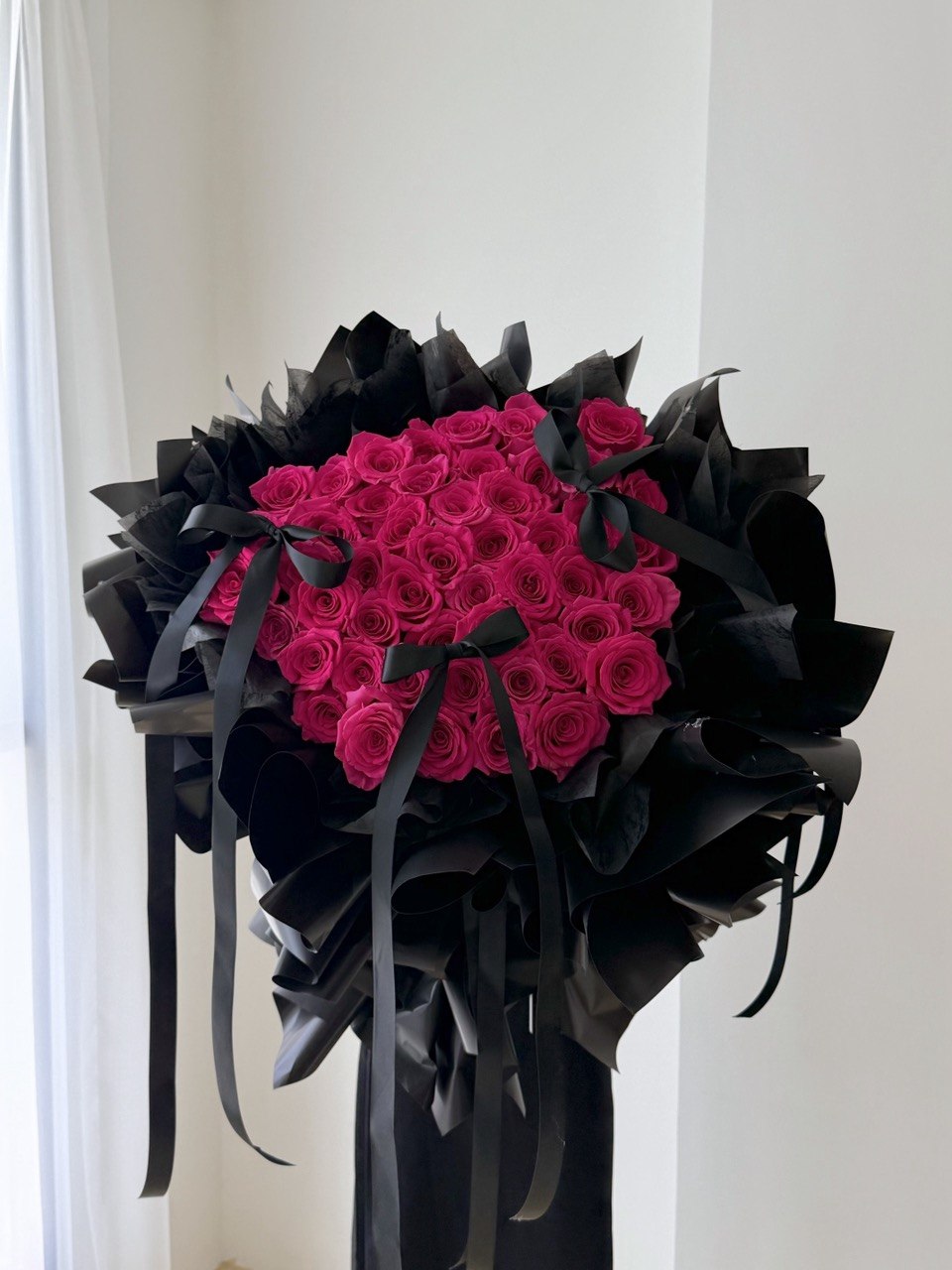 A large bouquet of 52 hot pink roses for proposal wrapped in dramatic black paper and tied with black ribbons, displayed indoors against a white wall.